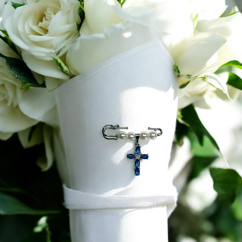 May include: A white bouquet of roses with a silver safety pin adorned with white pearls and a blue jeweled cross charm.