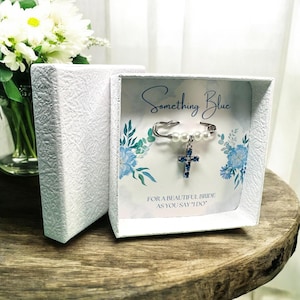 May include: A white box with a blue and white floral design. The box contains a silver safety pin with a blue cross charm and pearl beads. The box has the text "Something Blue" and "For a beautiful bride as you say 'I do'" printed on it.
