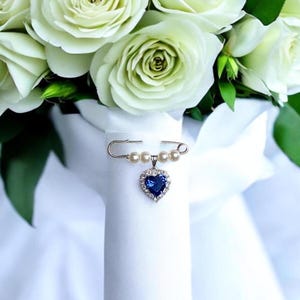 May include: A white bridal bouquet with a blue heart-shaped brooch with pearl accents. The brooch is attached to the bouquet with a safety pin.
