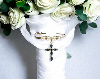 May include: A white bridal bouquet with a gold safety pin adorned with pearls and a blue sapphire cross charm.