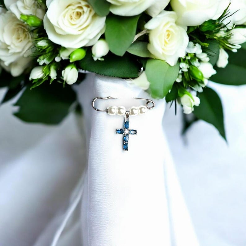 May include: A white bridal bouquet with a pearl and blue crystal cross charm attached to a safety pin.