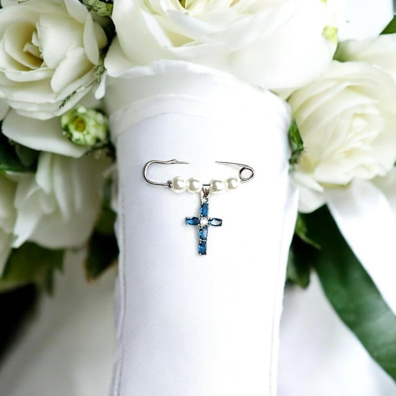May include: A white bridal bouquet with a silver safety pin adorned with white pearls and a blue jeweled cross charm.