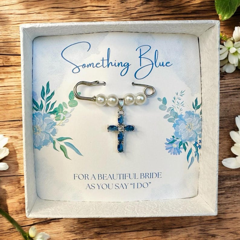 May include: A white box with a blue and white floral design. The box contains a silver safety pin with a blue cross charm and white pearl beads. The text "Something Blue" is printed on the box. The text "For a beautiful bride as you say "I do" is printed on the box.