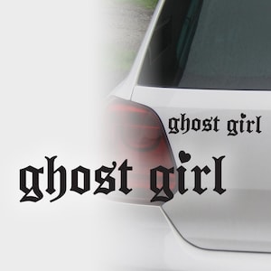 May include: A white car with a black "ghost girl" decal on the rear window. The decal is in a gothic font, with a small heart symbol. The car's taillight is visible.