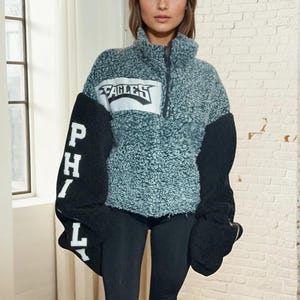 May include: A teal and black fleece jacket with a white "EAGLES" logo on the front and "PHILLY" on the sleeve. The jacket has a zip fastening and a stand-up collar.