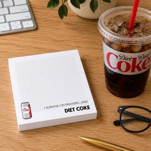 May include: A white rectangular notepad featuring a graphic of a Diet Coke can in the bottom left corner and the black printed text "I SURVIVE ON PRAYERS...AND DIET COKE" positioned to the right of the can illustration on the bottom section.