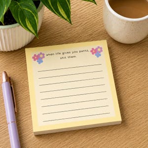 May include: A square notepad with a pale yellow background featuring a floral border of pink, blue, and purple flowers. The text reads, "when life gives you pants, shit them." Below the text are seven horizontal black dotted lines for writing.