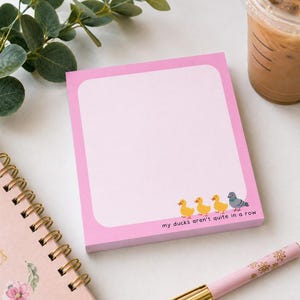 May include: A square notepad with a bright pink border and a large white center area. The bottom features three small yellow duck illustrations followed by a grey pigeon and the black text: my ducks aren't quite in a row.