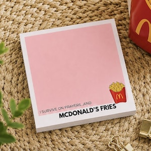 Puede incluir: Un bloc de notas cuadrado con fondo rosa claro que presenta una pequeña ilustración de un cartón rojo de patatas fritas doradas en la esquina inferior derecha. El texto dice, I SURVIVE ON PRAYERS...AND MCDONALD'S FRIES en letras negras.