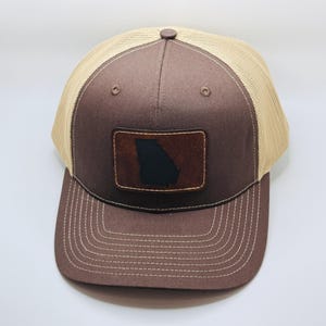 May include: A brown and tan trucker hat with a brown leather patch featuring the outline of the state of Georgia.
