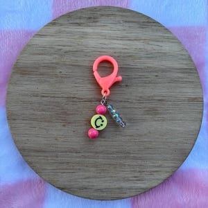 Puede incluir: Un llavero rosa neón con un mosquetón. El llavero presenta una cuenta amarilla con una "C" negra, dos cuentas rosas y una hebra de cuentas iridiscentes. El llavero está sobre una superficie de madera.