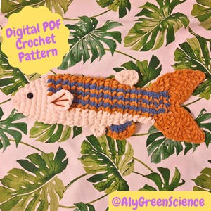 May include: A crocheted fish toy in white, orange, and blue, with a black eye and small fins. The fish is placed on a background of green tropical leaves. The image includes the text "Digital PDF Crochet Pattern" and "@AlyGreenScience".