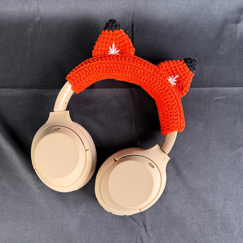 May include: Beige wireless headphones with a crocheted fox headband in orange and black.