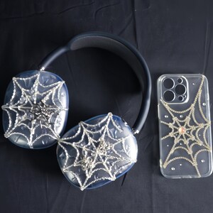 May include: A pair of black headphones with a spiderweb design and a clear phone case with a spiderweb design. Both items are covered in rhinestones.
