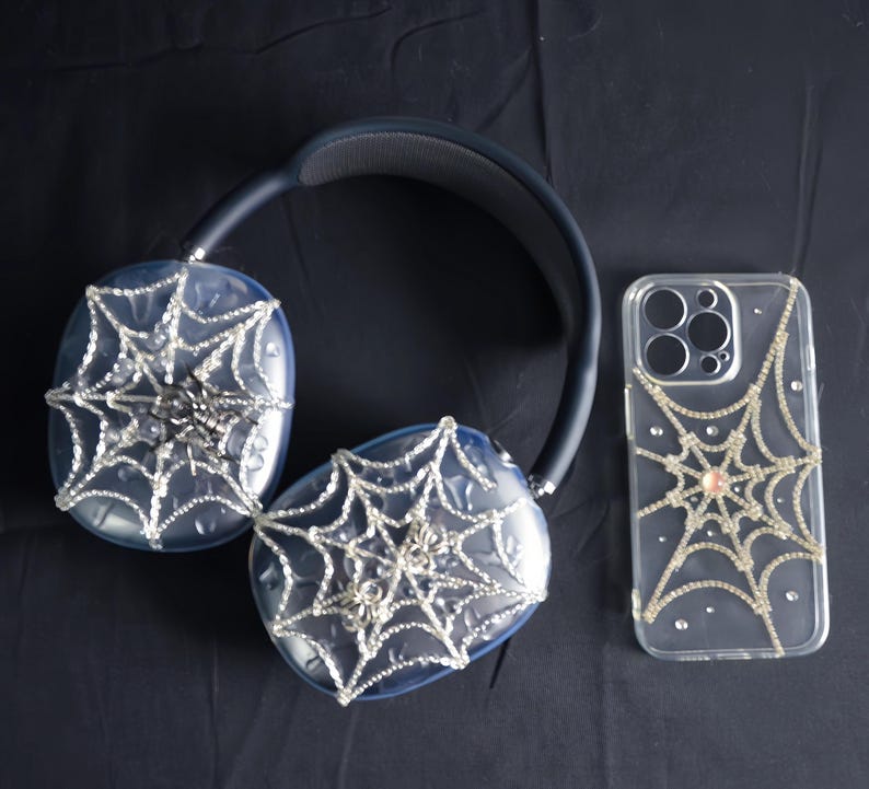 May include: A pair of black headphones with a spiderweb design and a clear phone case with a spiderweb design. Both items are covered in rhinestones.