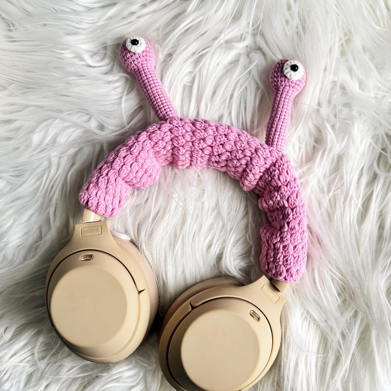 Op de afbeelding: Roze gehaakt hoofdband met oogjes, ontworpen om over koptelefoons te passen. Het hoofdband ligt op een wit, pluche oppervlak.