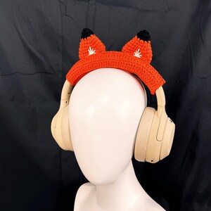May include: Orange crocheted headphone headband with fox ears. The ears are black with white accents. The headband is on a beige mannequin head.