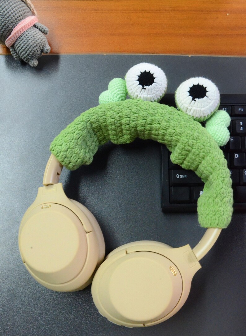 May include: Beige wireless headphones with a green crocheted headband featuring two white eyes with black pupils. The headband is shaped like a monster with a green crocheted body.