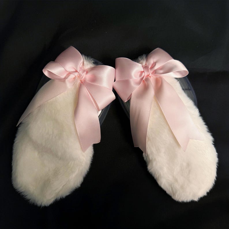 May include: A pair of white fluffy bunny ears with pink satin bows. The bows are tied in a knot at the center and have long tails.