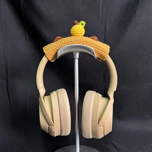 May include: Beige headphones with a crocheted brown and yellow headband with a small yellow pear on top. The headphones are on a metal stand.