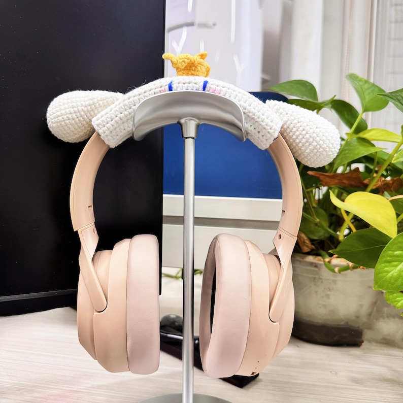 May include: Blush-colored headphones with a white crocheted ear cover and a small yellow crocheted crown. The headphones are displayed on a silver stand, with a green plant in the background. The ear cover has a cartoon-like design.