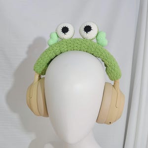 May include: Green crocheted frog headband with white eyes and black pupils, worn over a pair of beige headphones.