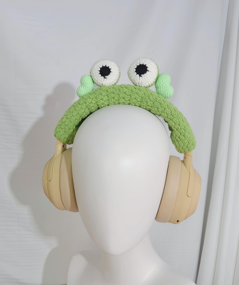 May include: Green crocheted frog headband with white eyes and black pupils, worn over a pair of beige headphones.