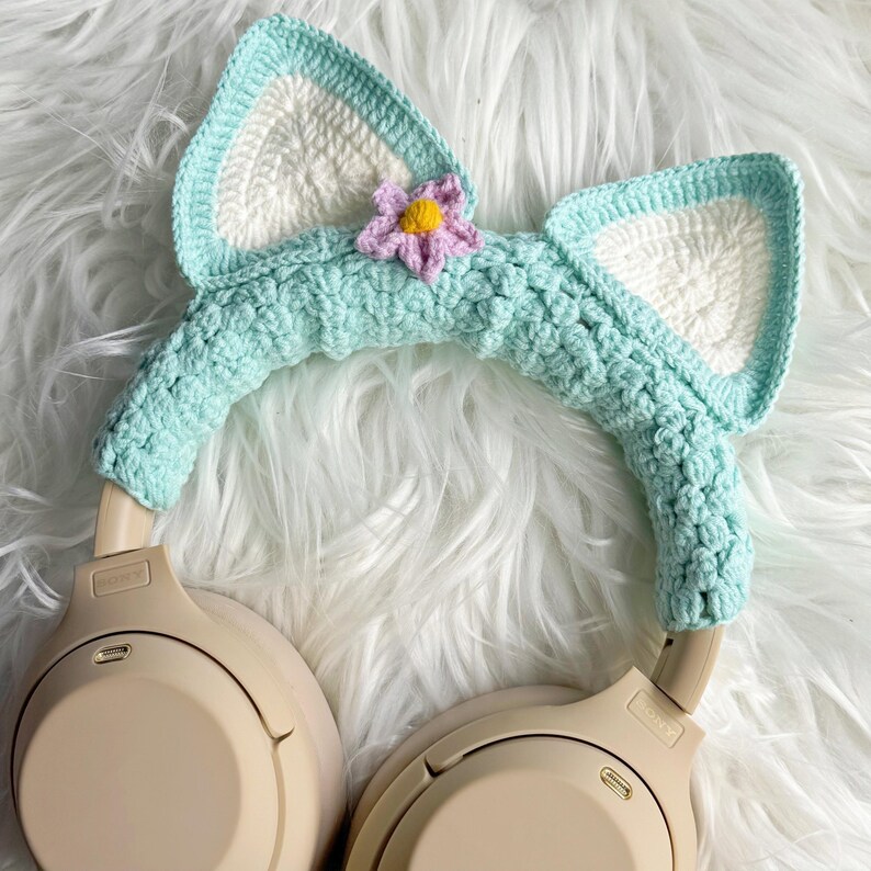 May include: Mint green crocheted cat ear headband with a pink flower accent, covering the headband of a pair of beige headphones.