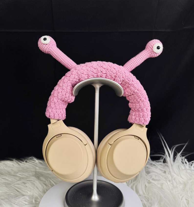 Op de afbeelding: Roze gehaakte hoofdtelefoonstandaard met twee wormachtige armen met ogen. De standaard houdt een paar beige hoofdtelefoons.