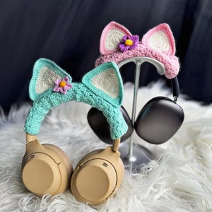 May include: Two pairs of crocheted cat ear headphone covers. One pair is pink with a purple flower and the other is light blue with a pink flower. The covers are on a pair of black headphones.