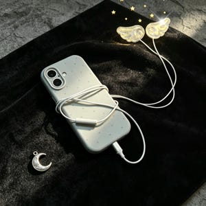 Può includere: Una custodia per smartphone grigio chiaro con un sottile motivo a cuori, dotata di un auricolare bianco con filo con due coperture traslucide e glitterate a forma di ala e un piccolo ciondolo a mezzaluna color argento.