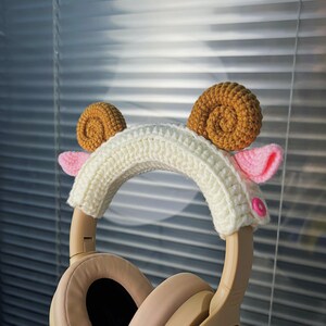 K&ouml;nnte beinhalten: Wei&szlig;er H&auml;kel-Stirnband mit braunen und rosa H&auml;kel-Widderh&ouml;rnern. Das Stirnband befindet sich auf einem beigefarbenen Kopfh&ouml;rer.
