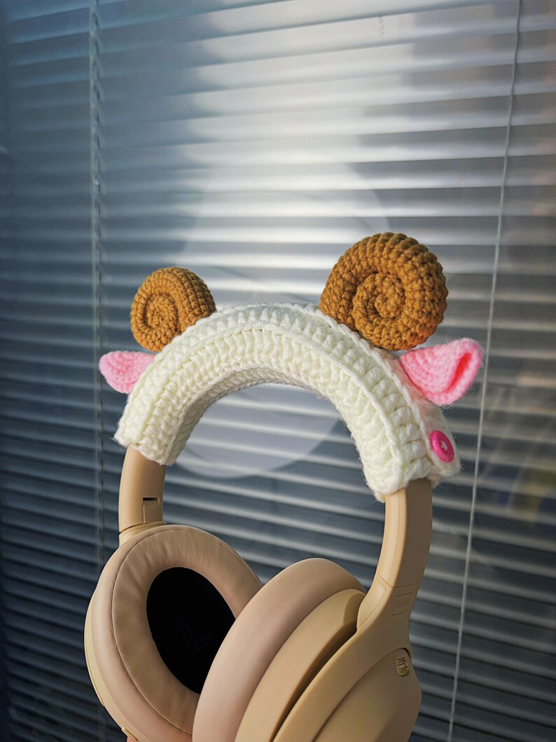 K&ouml;nnte beinhalten: Wei&szlig;er H&auml;kel-Stirnband mit braunen und rosa H&auml;kel-Widderh&ouml;rnern. Das Stirnband befindet sich auf einem beigefarbenen Kopfh&ouml;rer.