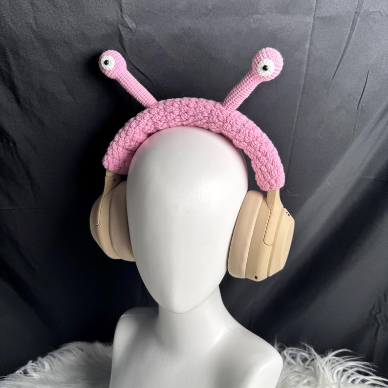 Op de afbeelding: Een roze gehaakt hoofdband met twee roze worm-achtige antennes met witte ogen. De hoofdband wordt gedragen over een beige koptelefoon.