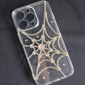 May include: A clear phone case with a silver rhinestone spiderweb design. The spiderweb has a pink rhinestone in the center.
