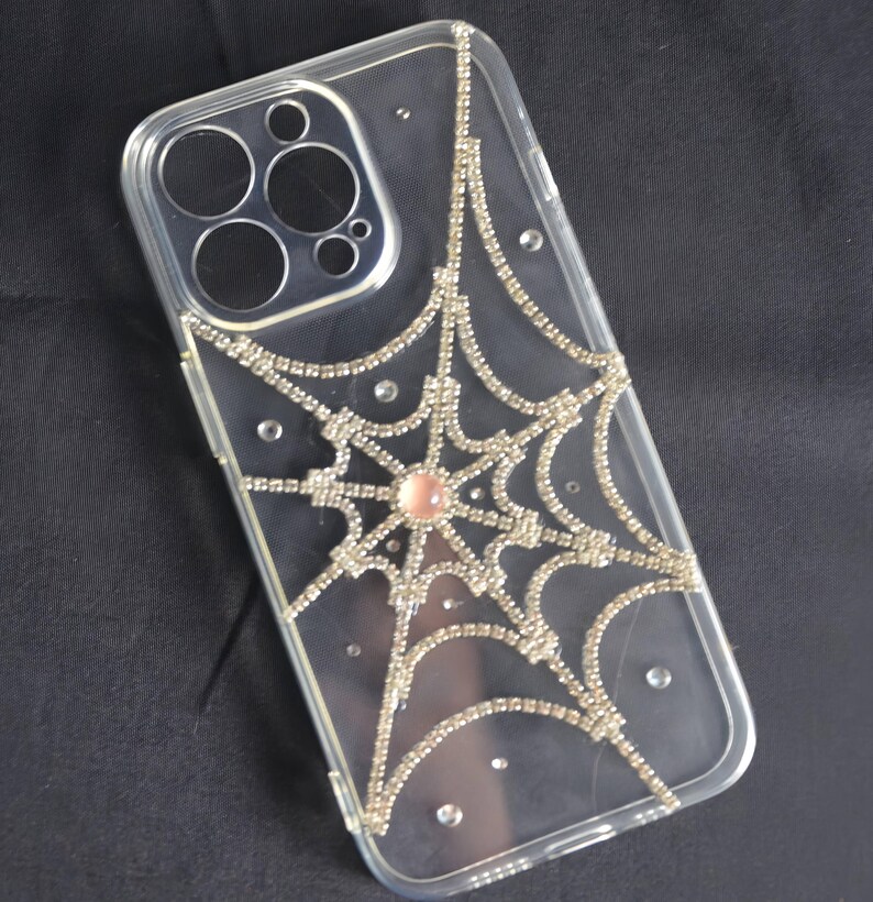 May include: A clear phone case with a silver rhinestone spiderweb design. The spiderweb has a pink rhinestone in the center.