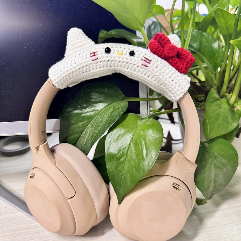 May include: Beige wireless headphones with a white crocheted cat ear headband with a red bow. The headband has embroidered eyes and a pink nose.