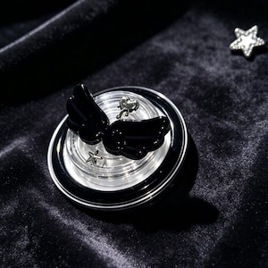 May include: A circular phone grip featuring a glossy black wing emblem centered on a clear, tiered base. The design includes a small silver-toned heart charm and a tiny silver star accent. A separate, detached silver star charm rests nearby.
