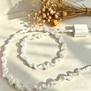 May include: A white charging cable encased in a ruffled, satin-like fabric sleeve featuring small, evenly spaced silver-toned embellishments. The cable is connected to a white power adapter and coiled into a circular shape on a light surface.