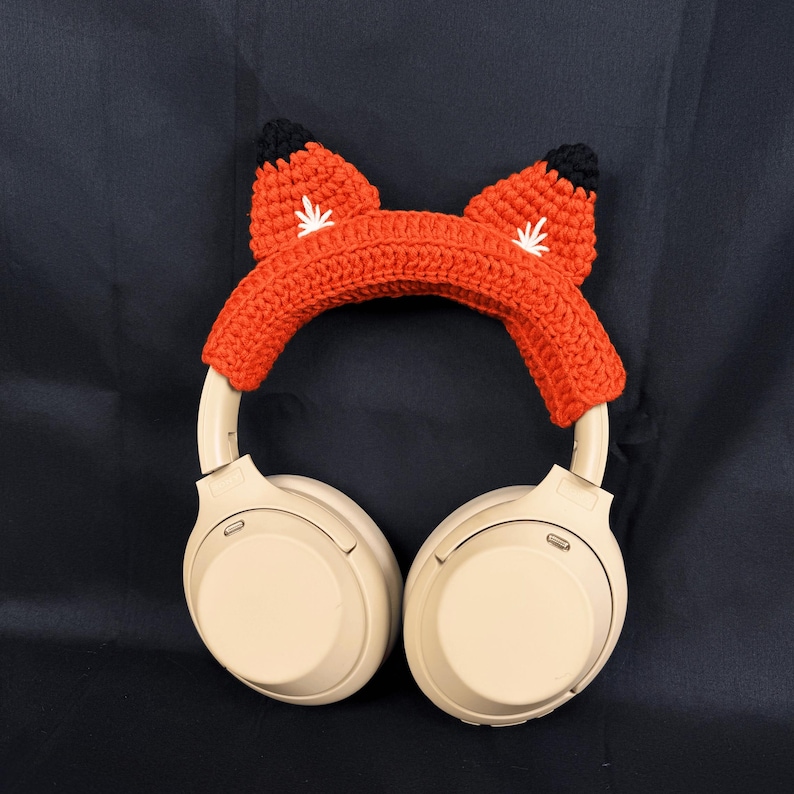 May include: Beige wireless headphones with a crocheted fox ear headband in orange and black.