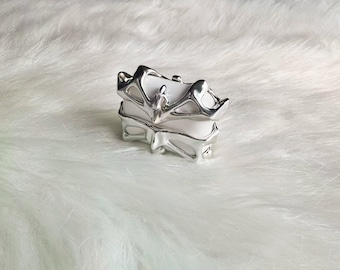 May include: A silver ring with an abstract, openwork design. The ring has a layered appearance, with a central element and a textured, organic shape. The ring is set against a white, fluffy background.