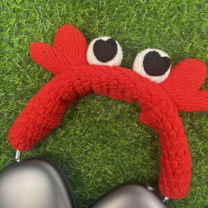 Op de afbeelding: Rode gebreide hoofdband in de vorm van een krab met witte ogen en zwarte pupillen. De hoofdband is ontworpen om over koptelefoons te passen.