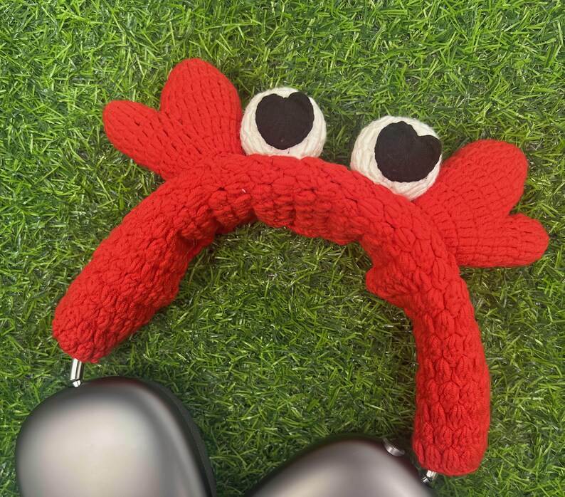Op de afbeelding: Rode gebreide hoofdband in de vorm van een krab met witte ogen en zwarte pupillen. De hoofdband is ontworpen om over koptelefoons te passen.