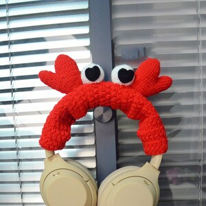 May include: Red crocheted crab headphone holder with white eyes. The holder is on a pair of beige over-ear headphones.