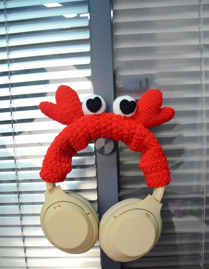 May include: Red crocheted crab headphone holder with white eyes. The holder is on a pair of beige over-ear headphones.