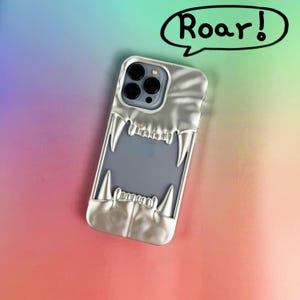 Op de afbeelding: Zilveren telefoonhoesje met een gestileerd ontwerp van een brullende dierenmond, met scherpe tanden. De hoes zit op een gradiënt achtergrond van roze, oranje, groen en blauw. Een tekstballon met het woord "Roar!" staat boven de telefoon.