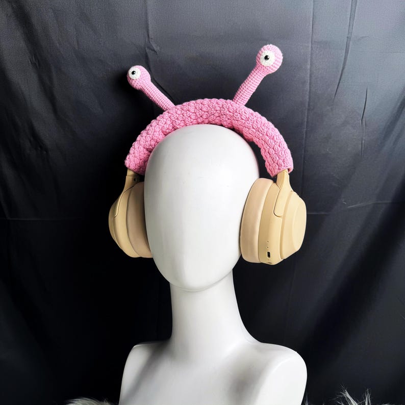 Op de afbeelding: Een roze gehaakt hoofdband met twee roze ogen erop, gedragen over een beige koptelefoon.