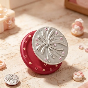 May include: A round, deep red compact mirror featuring a silver-toned lid embossed with a floral firework pattern and accented with small pink crystals. The word "Ceseclest" is engraved on the lid. A smaller matching circular piece sits nearby.