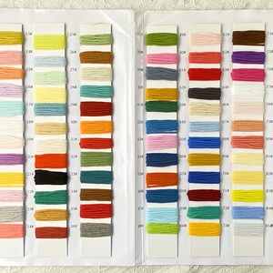 Op de afbeelding: Een kleurenkaart met rijen van kleine garenstalen in verschillende kleuren. De kaart is verdeeld in secties met nummers en letters. De garenstalen zijn in een rasterpatroon gerangschikt.