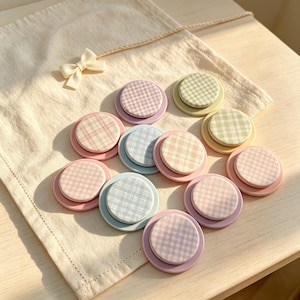 May include: A collection of eleven circular phone grips featuring pastel gingham fabric patterns in shades of pink, blue, green, and purple, each mounted on a matching solid-colored plastic base, arranged on a cream-colored fabric pouch.
